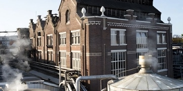 nos_brasseries_tuiles Bâtiment de style industriel avec des tuyaux visibles à l'extérieur, de la vapeur s'échappant des cheminées, représentant une brasserie.