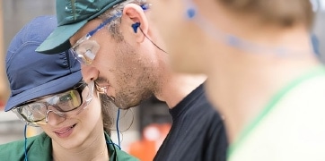 nos_valeurs_tuiles Trois employés portant des lunettes de protection et des bouchons d'oreilles discutent dans une usine, mettant l'accent sur la sécurité au travail.