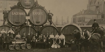 notre_histoire_tuiles Photographie vintage en noir et blanc montrant des ouvriers devant de grandes barriques de bière marquées 'Lagerbier Export', représentant une ancienne brasserie.